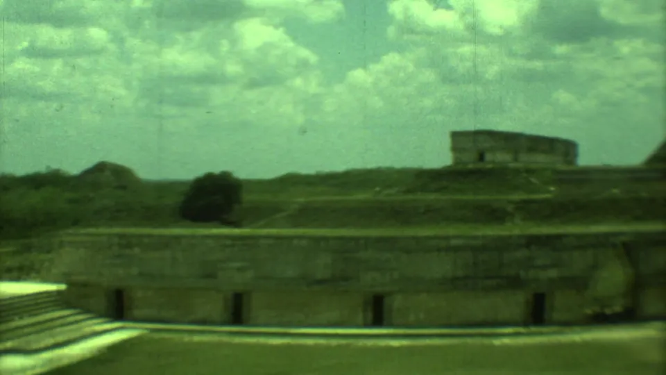 1973 8mm view across a Yucatan archaeological courtyard toward a distant stone structure and surrounding greenery.