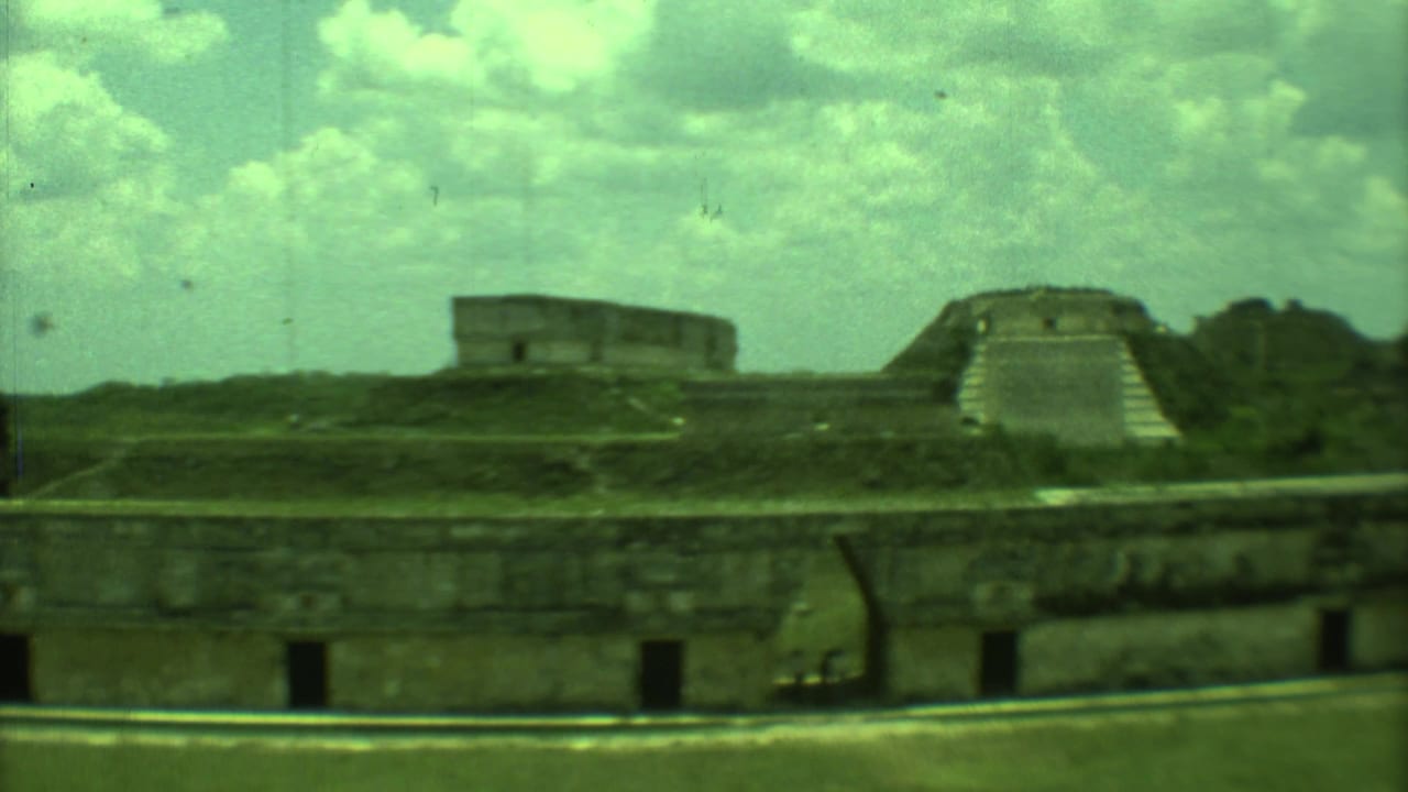 1973 Yucatan 8mm view of ancient stone ruins with a stepped temple form, grassy mounds, and a low wall in front.