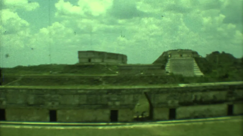 1973 Yucatan 8mm view of ancient stone ruins with a stepped temple form, grassy mounds, and a low wall in front.