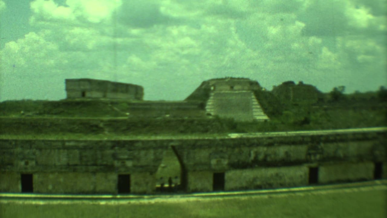 Wide view of stone temple ruins with grassy slopes and a low wall in front.