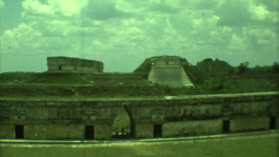 1973 Yucatan home-movie frame showing a wide archaeological-site view with temple ruins, grass, and cloud cover.