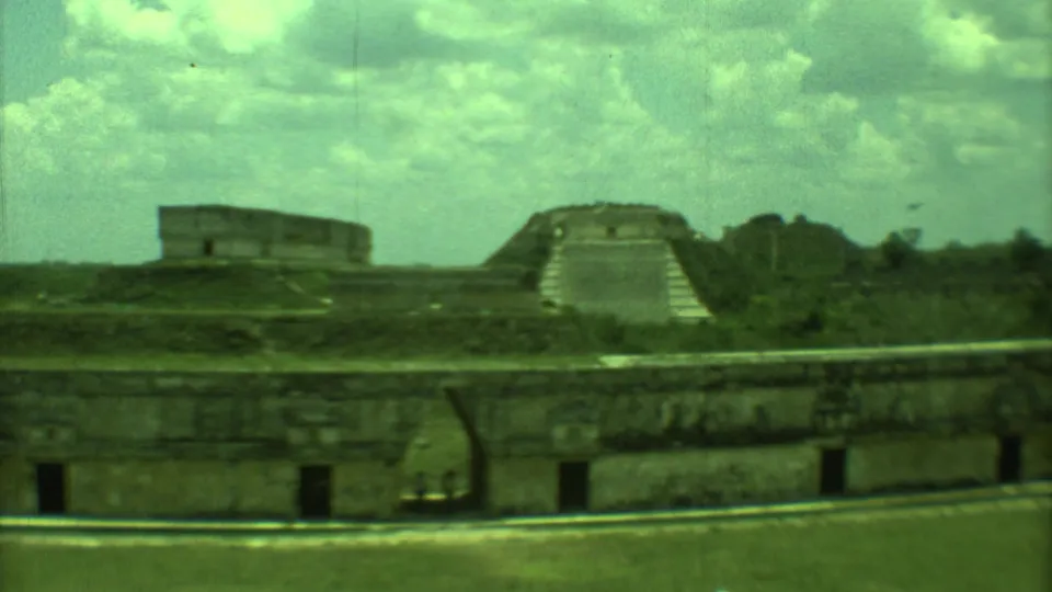1973 Yucatan 8mm frame with low stone wall openings, distant ruins, a stepped structure, and cloudy sky.