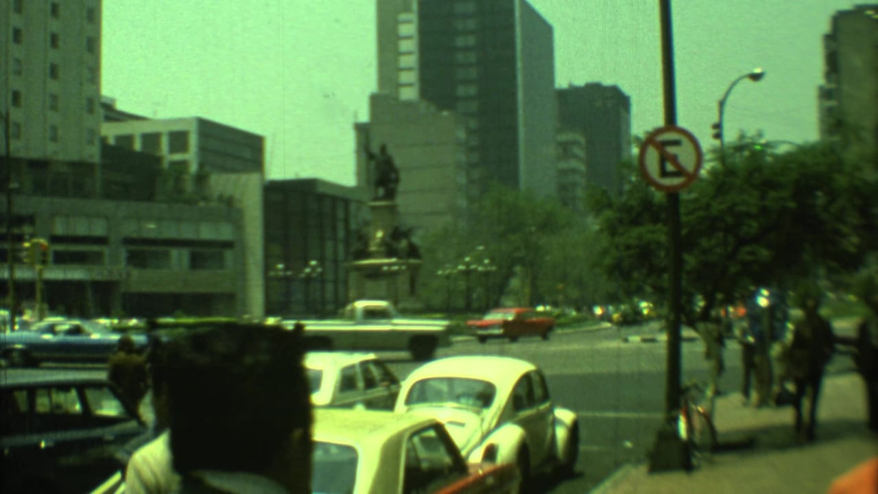 1973 Yucatan, Mexico 8mm frame showing traffic near a monument plaza with pedestrians and modern buildings beyond.