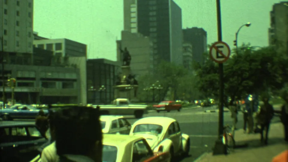 1973 Yucatan, Mexico 8mm frame showing traffic near a monument plaza with pedestrians and modern buildings beyond.