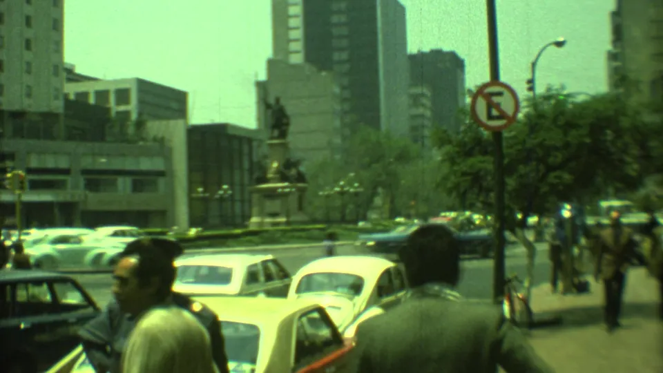 Handheld 1973 Yucatan, Mexico street view with Beetles, pedestrians, traffic signs, and a statue in a central plaza.