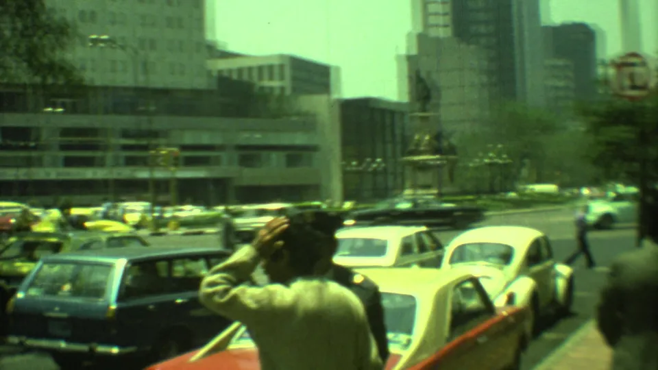 1973 Yucatan, Mexico 8mm frame of traffic around a square, with a foreground pedestrian and office buildings visible.