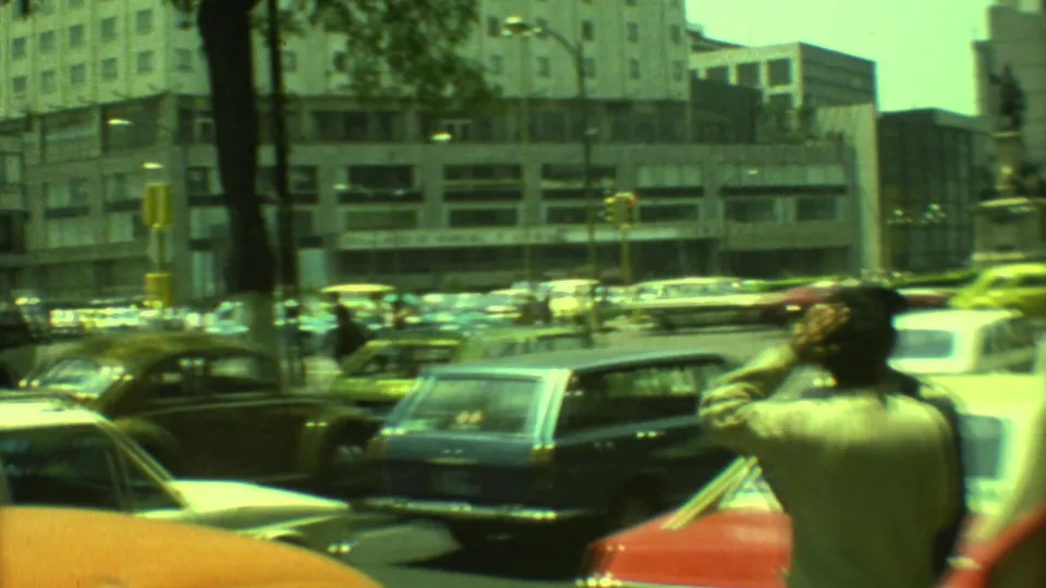 1973 Yucatan, Mexico street scene with a dark station wagon, Beetles, pedestrians, and plaza buildings under green-shifted 8mm color.