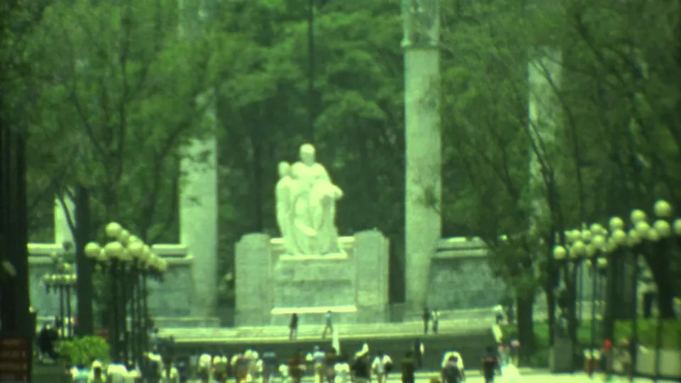 Closer 1973 8mm view of the monument area in Yucatan, with a statue, tall columns, iron fencing, trees, and distant visitors.