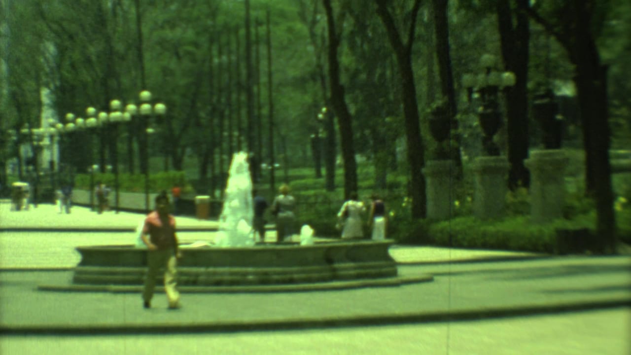 1973 Yucatan, Mexico home-movie view of a tree-lined urban plaza with a fountain, pedestrians, lamps, and city buildings.