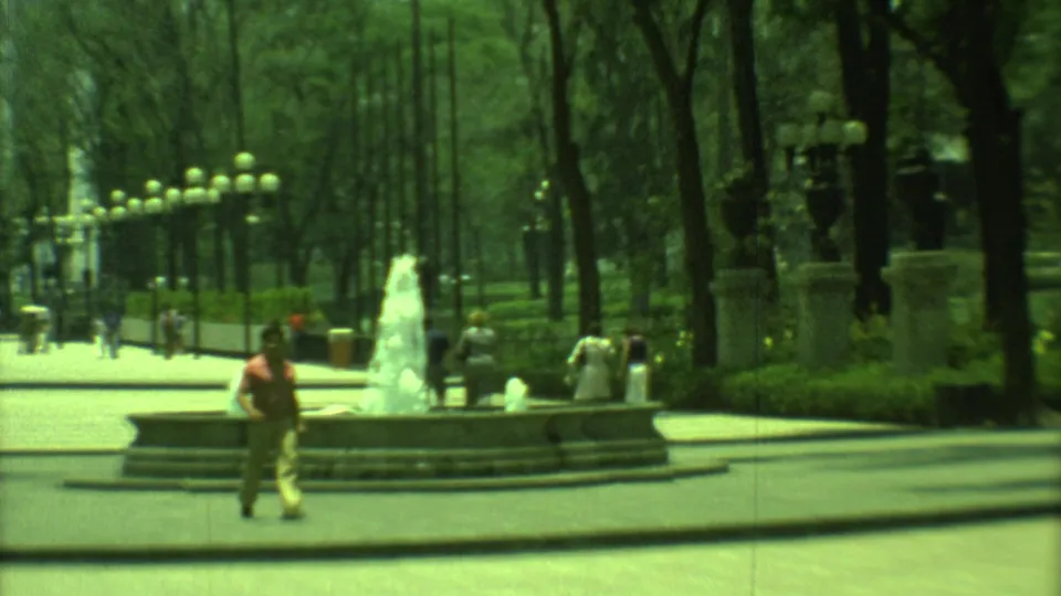 1973 Yucatan, Mexico home-movie view of a tree-lined urban plaza with a fountain, pedestrians, lamps, and city buildings.