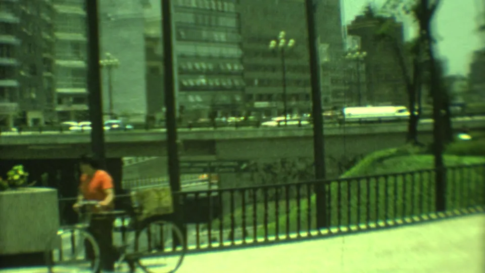 1973 Yucatan, Mexico street view with a cyclist by a railing, an overpass carrying cars, and modern city buildings in the background.