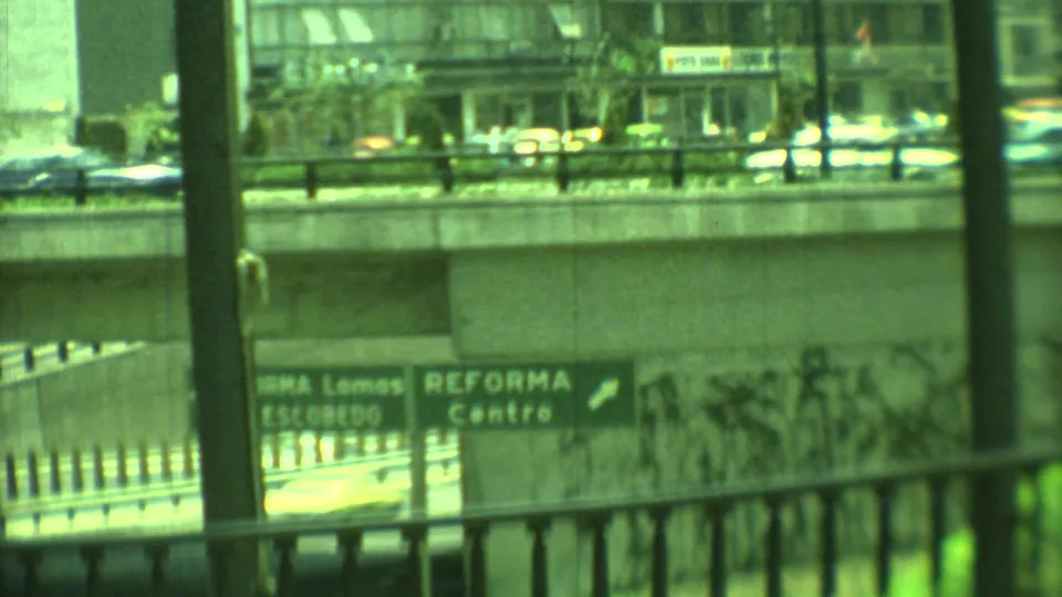 1973 Yucatan, Mexico home-movie frame looking past dark railings toward a concrete overpass, directional signs, traffic, and urban buildings.
