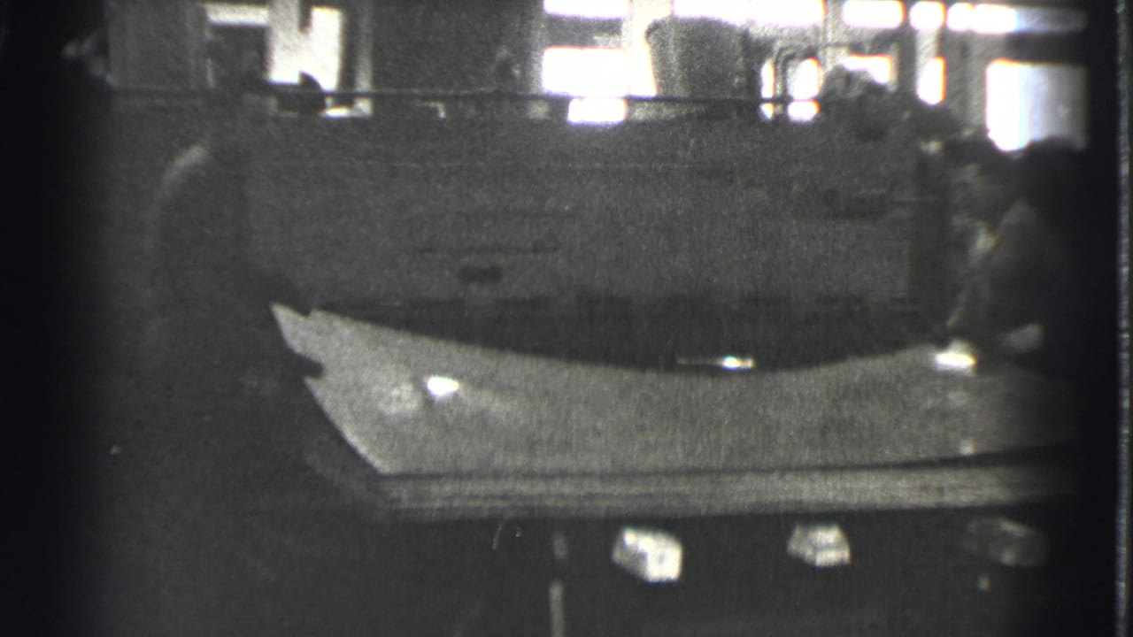 Two workers stand near a large machine while a flat sheet rests across the foreground.