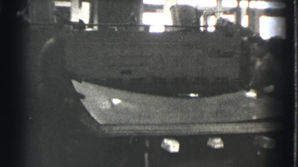 In a Cleveland factory scene from 1939, workers handle a broad flat object beside a large industrial machine.