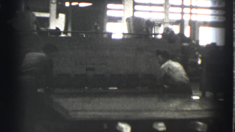 In a 1939 Cleveland factory interior, two adult workers appear near a large sheet metal press beneath bright windows.