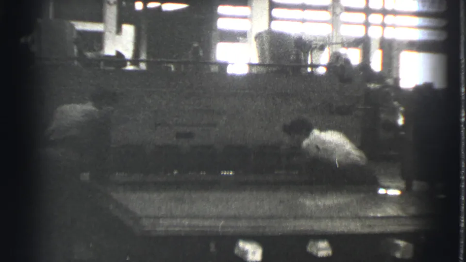 A worker in a light shirt leans beside a large sheet metal press in the Cleveland factory scene, with another figure partly visible.