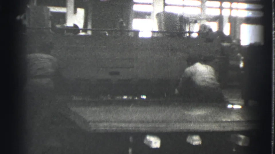 A worker seen from behind stands near the large metal press, with factory windows and another figure in the background.