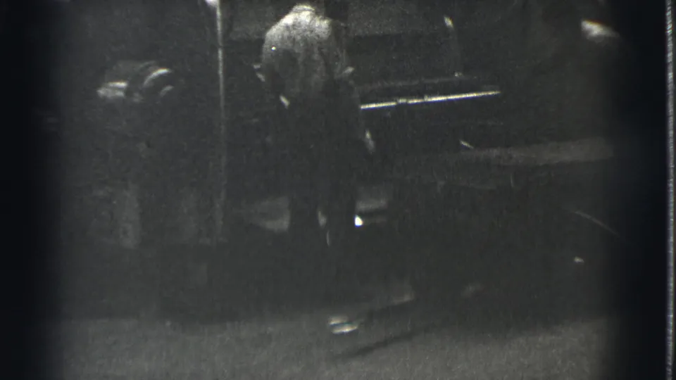 A worker in the Cleveland industrial scene stands close to a large machine, partly obscured by darkness and grain.
