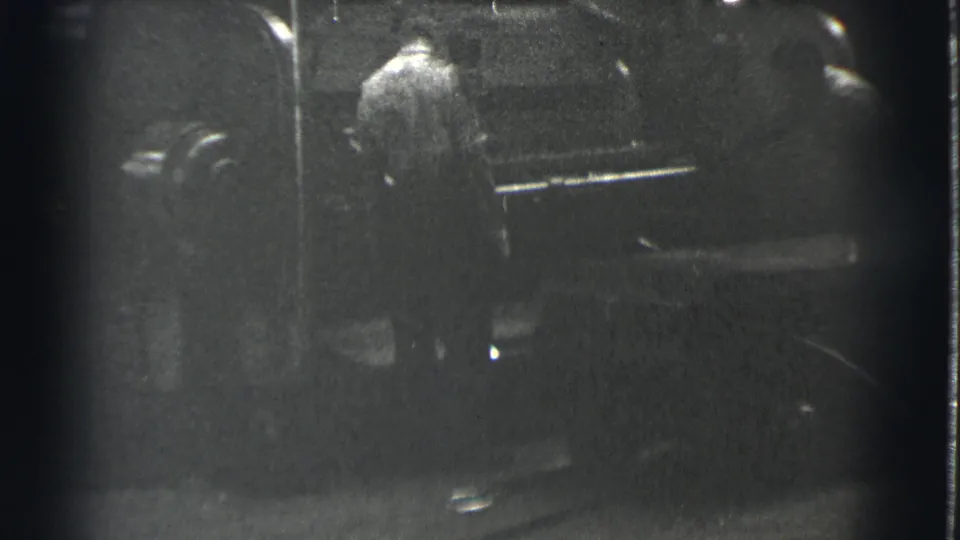 Black-and-white 8mm frame shows a worker near heavy equipment, with low exposure and a small bright reflection on the floor.