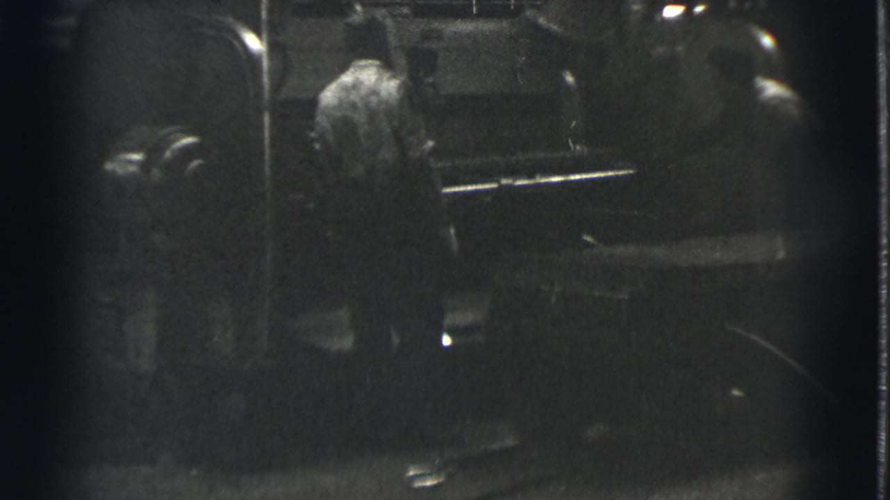 In 1939 Cleveland home-movie footage, two adult men appear beside a large machine in a dim industrial setting.