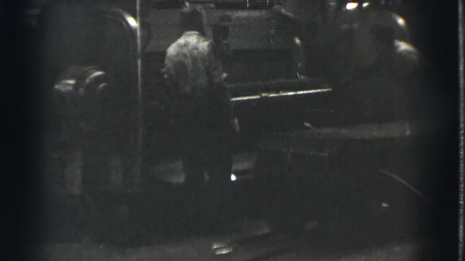 In 1939 Cleveland home-movie footage, two adult men appear beside a large machine in a dim industrial setting.