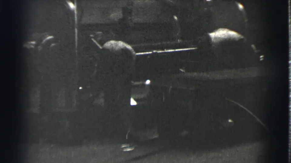 One adult man bends or leans near the machine while another figure stands nearby in the dim Cleveland industrial setting.
