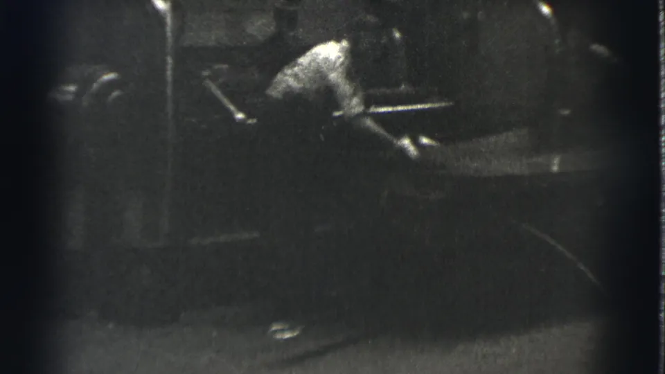 In 1939 Cleveland home-movie footage, a worker in a light shirt leans toward heavy machinery in a dim shop setting.