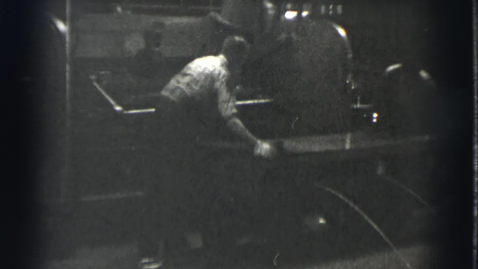 A man works close to large rounded machinery, with soft focus and heavy grain from the 8mm black-and-white scan.