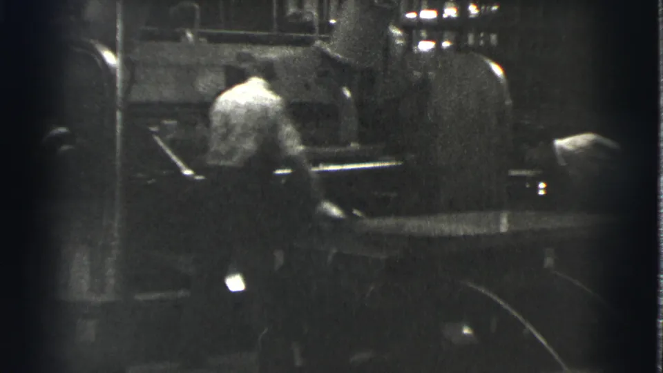 A worker in a light shirt stands by heavy machinery; a bright indistinct form at the right edge may be another person or object.