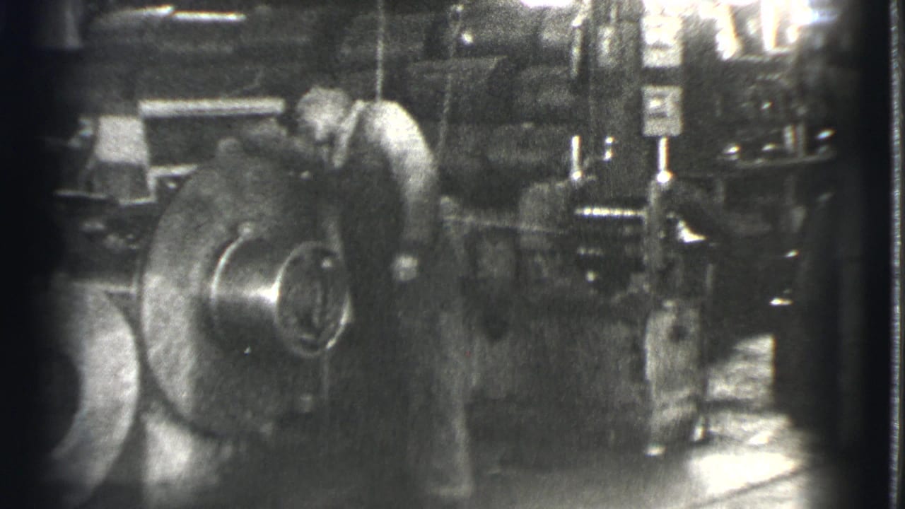 Worker bends over a large spinning metal machine inside a dim factory.