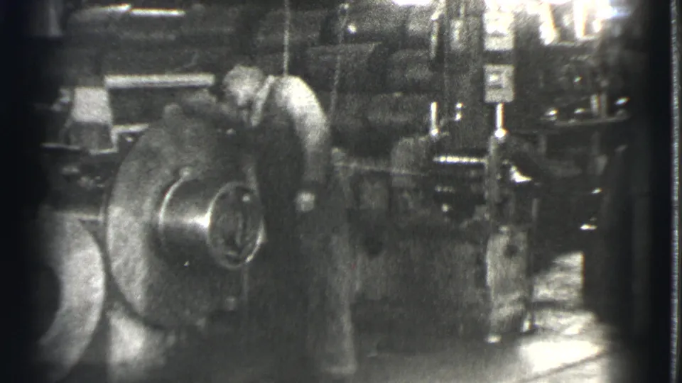 In Cleveland, Ohio in 1939, a man tends a large rotating industrial component in a dark factory space.