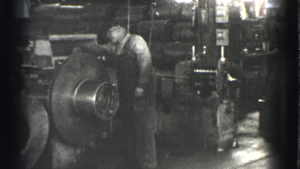 The frame shows a man leaning over heavy industrial equipment, with a large circular machine part filling the foreground.