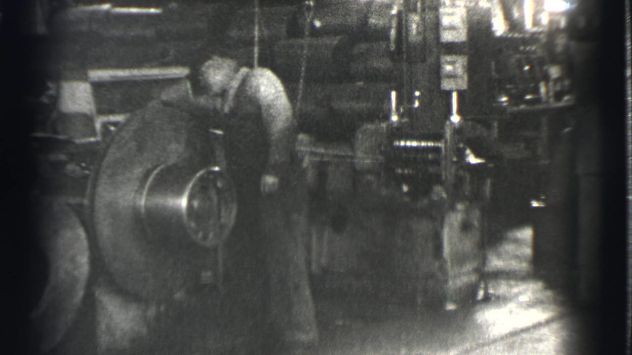 In 1939 Cleveland factory footage, a man in work clothes bends toward a large round machine amid gauges and metal equipment.