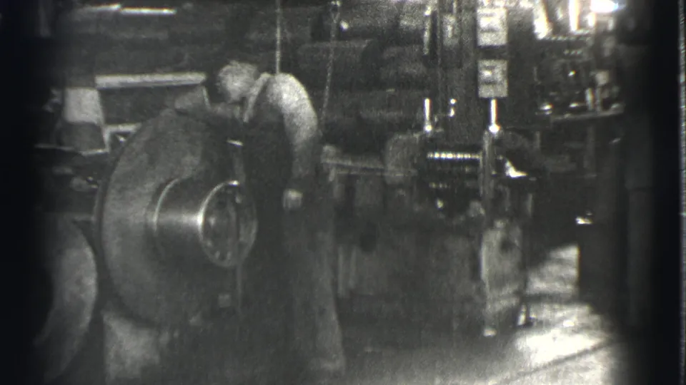 In 1939 Cleveland factory footage, a man in work clothes bends toward a large round machine amid gauges and metal equipment.