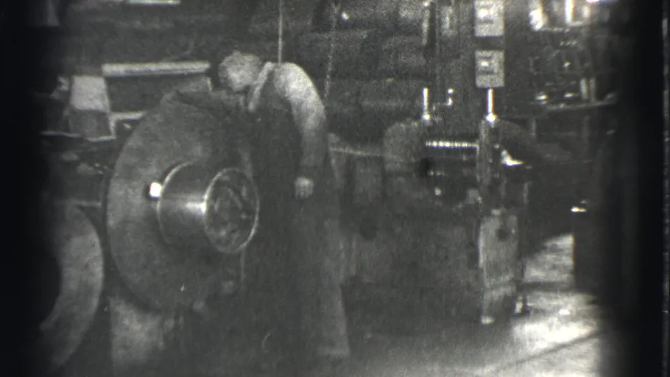 The worker remains beside the large industrial machine, with gauges, pipes, and heavy equipment visible in the Cleveland factory interior.