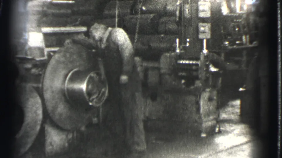 A man leans toward the industrial machine as black-and-white 8mm grain and edge flare mark the factory shot.