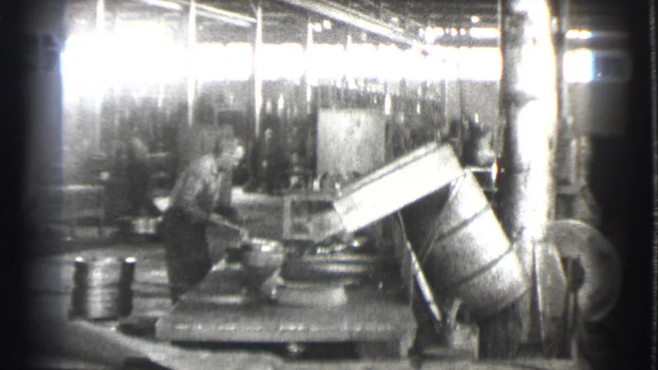 In 1939 Cleveland factory footage, a worker stands near heavy machinery, an angled metal component, ducting, and bright upper windows.