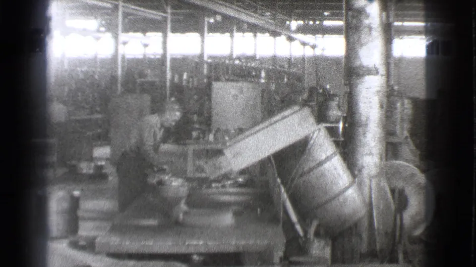 A worker appears to operate or inspect heavy equipment inside the Cleveland factory, with machinery, columns, and bright window light behind him.
