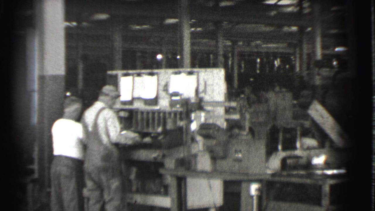 Two men stand at a large machine inside a dim factory workshop.