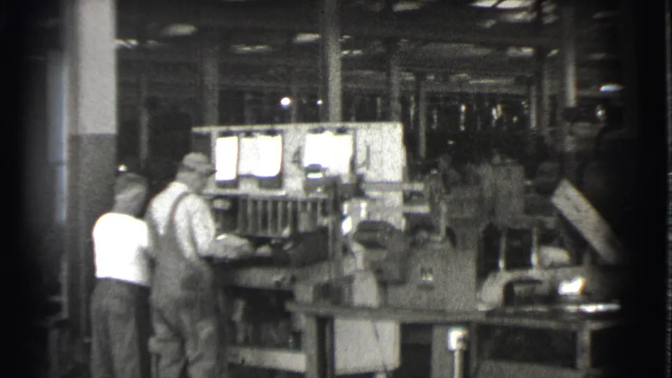 In 1939 Cleveland footage, two men in work clothes stand beside a large industrial machine in a dim factory interior.
