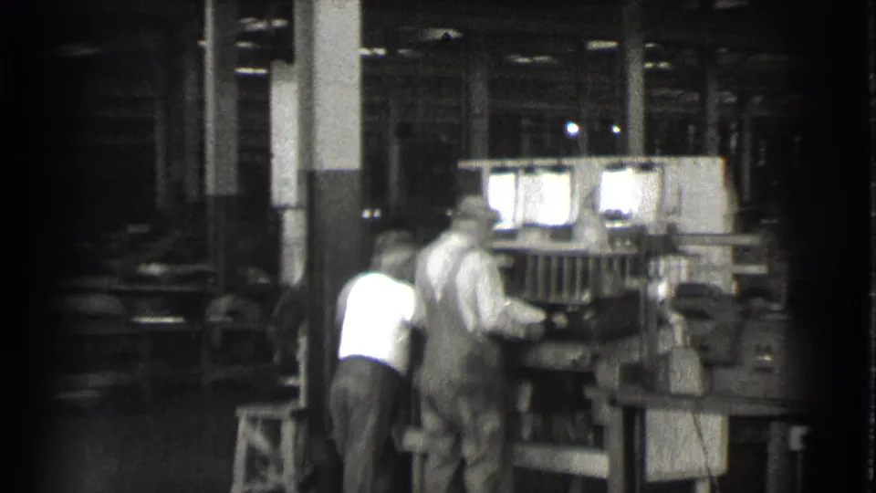 Two men in work clothes lean toward a large machine during a handheld pan inside a 1939 Cleveland factory setting.