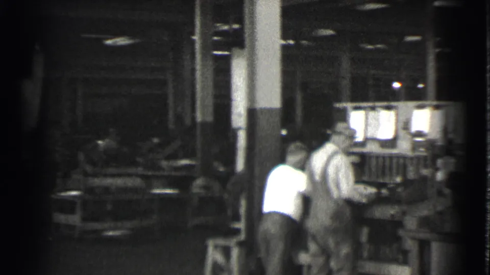A man bends at the machinery while another worker stands nearby in the Cleveland factory footage from 1939.