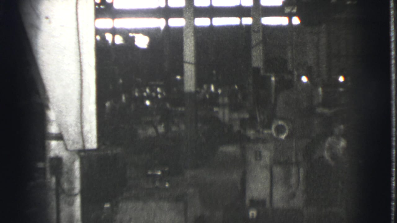 Dim factory interior with machinery, columns, and bright high windows.