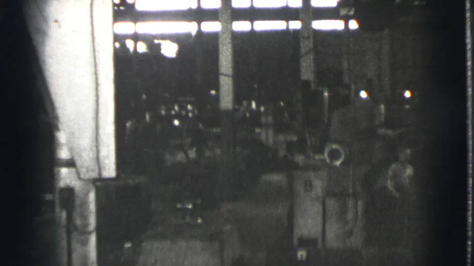 1939 Cleveland factory interior with large machinery, vertical columns, and silhouetted workers beneath bright high windows.