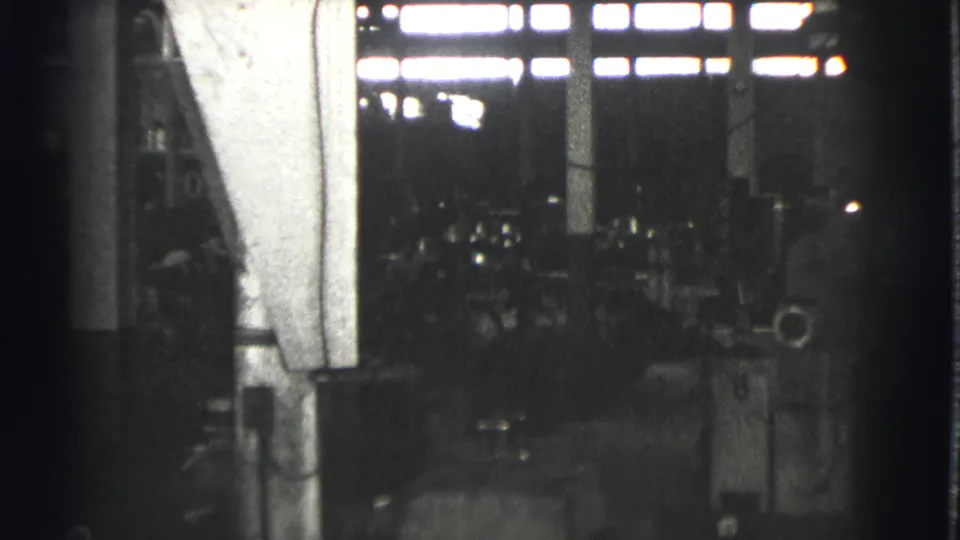 A dim Cleveland factory floor appears with heavy equipment, high window light, and workers partly obscured by machinery.