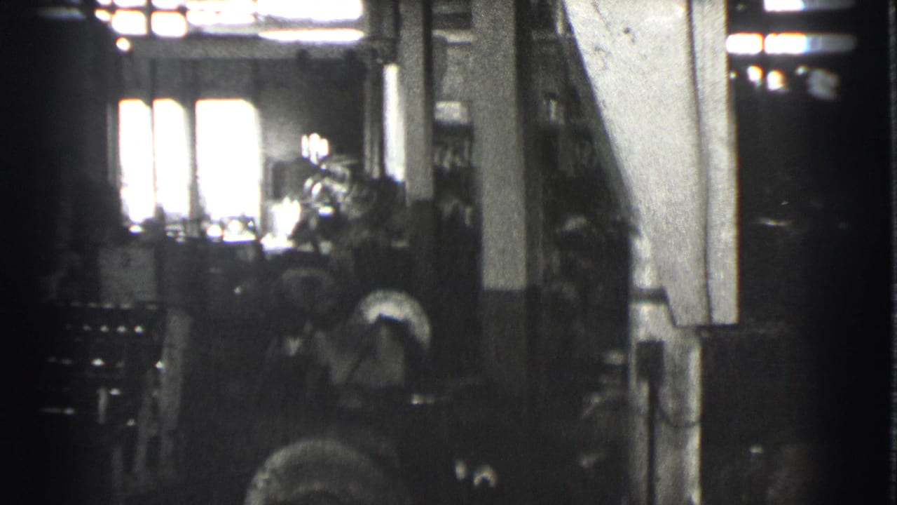 1939 Cleveland factory interior showing a worker beside heavy machinery, with high windows and columns visible in soft black-and-white 8mm footage.