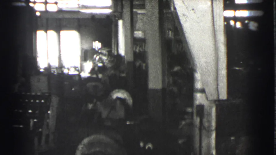 1939 Cleveland factory interior showing a worker beside heavy machinery, with high windows and columns visible in soft black-and-white 8mm footage.
