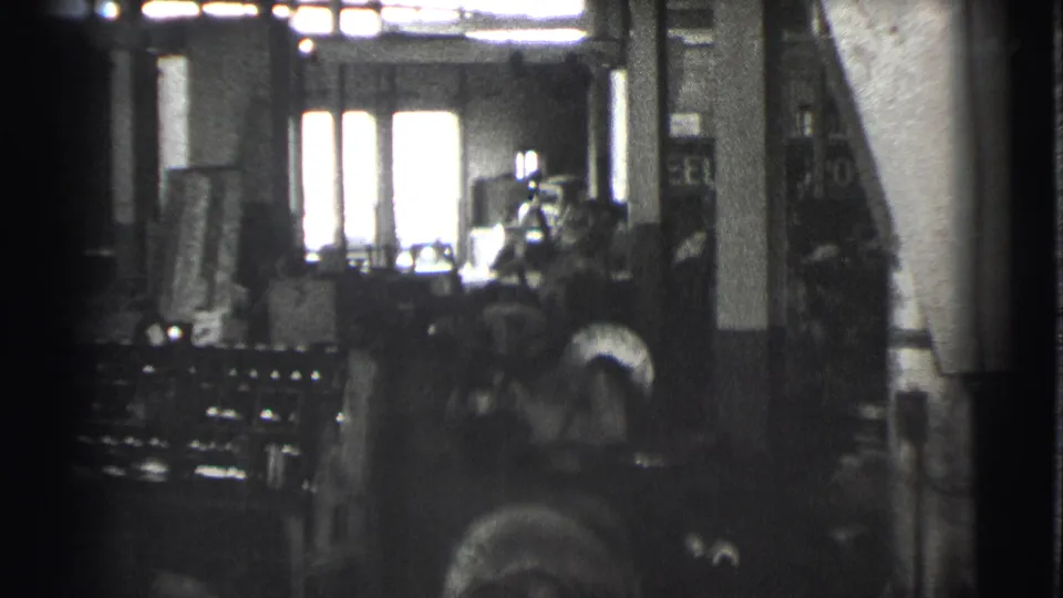 A black-and-white 8mm view of the Cleveland factory floor, with a worker near machinery and bright window light behind the equipment.
