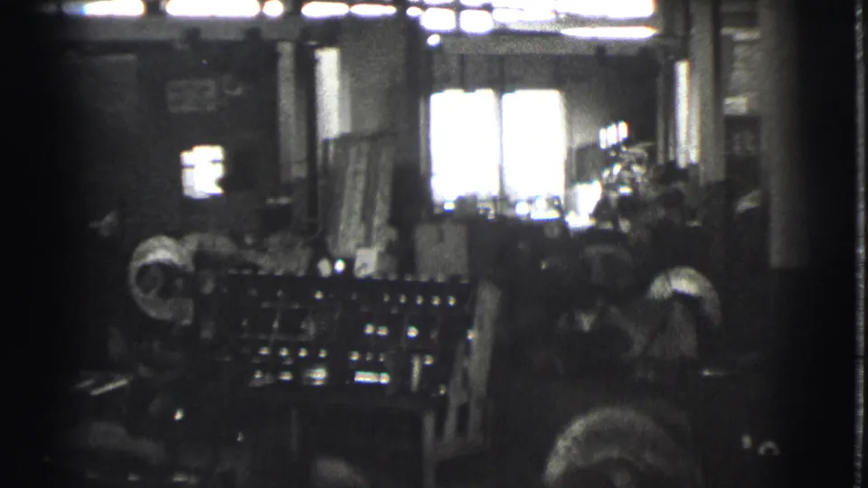 Two or more workers appear around factory equipment in the 1939 Cleveland interior, with benches, machinery, and strong window glare visible.