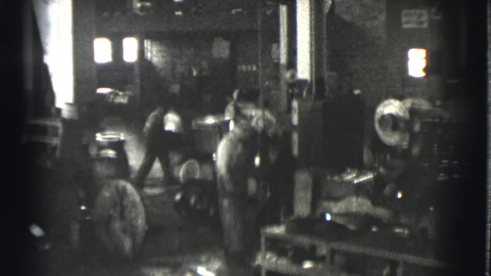 1939 Cleveland workers are seen around heavy machinery, benches, and containers in a grainy factory interior.